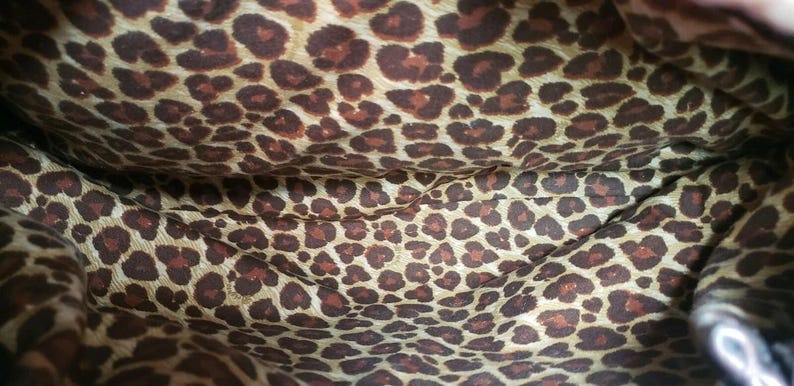 May include: Interior of a bag with a leopard print pattern. The fabric is a light tan color with dark brown spots. The pattern is repeated throughout the lining of the bag.
