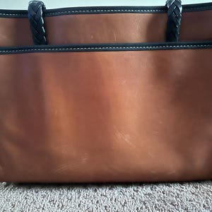 May include: A brown leather tote bag with black trim and braided handles. The bag has a rectangular shape with a large main compartment and an exterior pocket. White stitching accents the black trim.