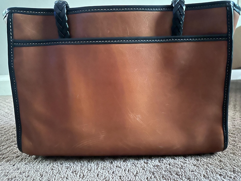 May include: A brown leather tote bag with black trim and braided handles. The bag has a rectangular shape with a large main compartment and an exterior pocket. White stitching accents the black trim.