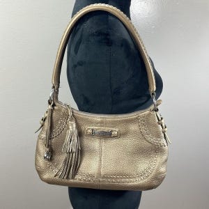 May include: A metallic gold shoulder bag with a textured finish. The bag features a braided handle, decorative stitching, and a tassel. A silver-toned metal bar is centered on the front. The bag is displayed on a black mannequin.