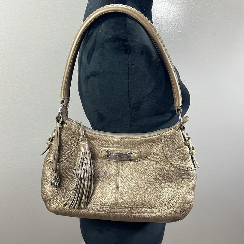 May include: A metallic gold shoulder bag with a textured finish. The bag features a braided handle, decorative stitching, and a tassel. A silver-toned metal bar is centered on the front. The bag is displayed on a black mannequin.