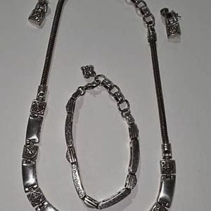 Puede incluir: Un conjunto de joyas plateadas que incluye un collar, una pulsera y un par de pendientes. El collar y la pulsera tienen eslabones rectangulares con detalles decorativos. Los pendientes son peque&ntilde;os, a juego con el dise&ntilde;o del collar y la pulsera.