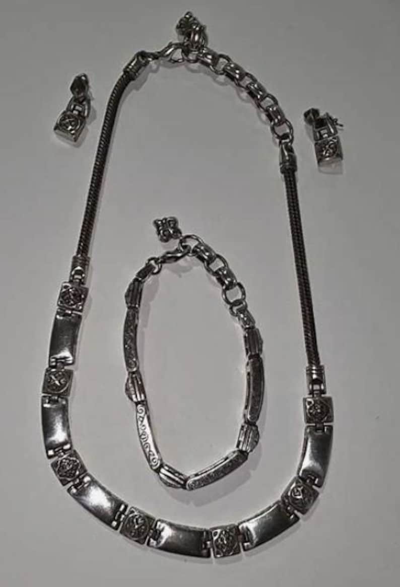 Puede incluir: Un conjunto de joyas plateadas que incluye un collar, una pulsera y un par de pendientes. El collar y la pulsera tienen eslabones rectangulares con detalles decorativos. Los pendientes son peque&ntilde;os, a juego con el dise&ntilde;o del collar y la pulsera.