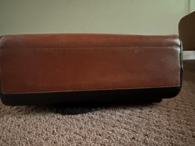 May include: A brown leather handbag with a black base. The bag has a rectangular shape with a stitched seam detail. The leather has a smooth texture and a rich, warm color.