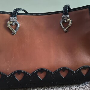 May include: A brown leather tote bag with black trim and braided handles. The bag features heart-shaped cutouts along the bottom edge and decorative heart-shaped clasps. The bag has a structured design and a polished finish.