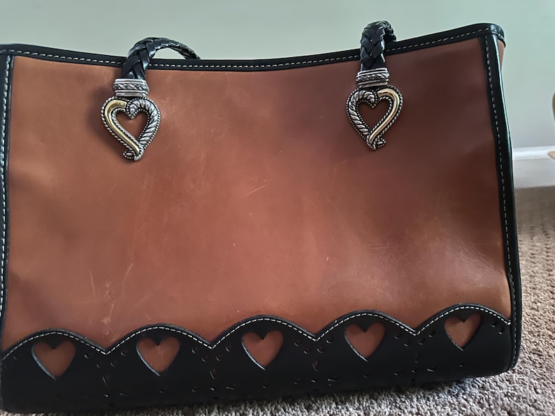 May include: A brown leather tote bag with black trim and braided handles. The bag features heart-shaped cutouts along the bottom edge and decorative heart-shaped clasps. The bag has a structured design and a polished finish.