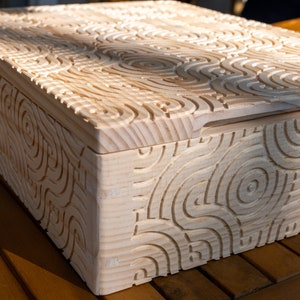 May include: A wooden box with a carved, intricate pattern on the lid and sides. The box is made of light wood and has a natural finish.