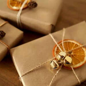 Pode incluir: Três presentes embrulhados em papel castanho com barbante e elementos decorativos. Um presente tem uma fatia de laranja seca e sinos dourados, outro tem uma fatia de laranja seca e uma pinha, e o terceiro tem uma pinha.
