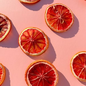 May include: Overhead shot of several dried blood orange slices arranged on a light pink background. The orange slices are a vibrant red-orange color with a lighter orange center and a tan rind. The image is well-lit, with shadows.