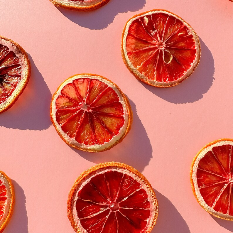May include: Overhead shot of several dried blood orange slices arranged on a light pink background. The orange slices are a vibrant red-orange color with a lighter orange center and a tan rind. The image is well-lit, with shadows.