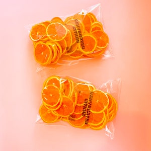 May include: Two clear plastic bags filled with bright orange dehydrated citrus slices, arranged on a light pink background. The bags have a printed label that reads "DEHYDRATED CITRUS". The slices are round and evenly cut.