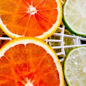 May include: Close-up of sliced oranges and limes. The oranges are a vibrant orange color with a white rind, and the limes are green. The fruit is arranged on a white grid, showcasing the juicy interior of the citrus.