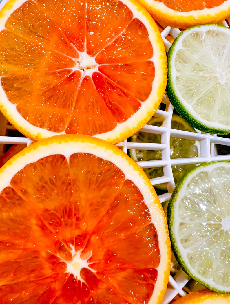 May include: Close-up of sliced oranges and limes. The oranges are a vibrant orange color with a white rind, and the limes are green. The fruit is arranged on a white grid, showcasing the juicy interior of the citrus.