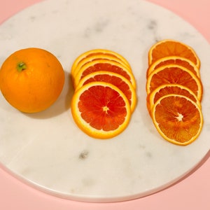 May include: An orange fruit and several slices of oranges are arranged on a white marble surface against a pink background. The orange slices vary in appearance, with some appearing fresh and others dried. The image highlights the vibrant colors and textures of the citrus fruit.