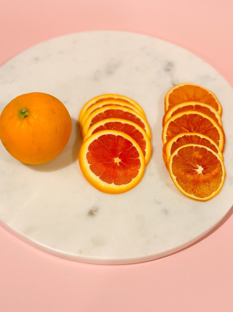 May include: An orange fruit and several slices of oranges are arranged on a white marble surface against a pink background. The orange slices vary in appearance, with some appearing fresh and others dried. The image highlights the vibrant colors and textures of the citrus fruit.