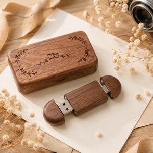 Puede incluir: Una caja rectangular de madera con tapa abatible que presenta un diseño grabado con láser de dos pequeños corazones rodeados de enredaderas. Junto a ella hay una memoria USB de madera a juego con una tapa extraíble.