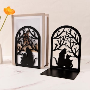 Könnte beinhalten: Zwei schwarze Katzen-Buchstützen aus Metall. Jede Buchstütze zeigt eine Silhouette von zwei Katzen unter einem Astmuster. Die Buchstützen stehen auf einer weißen Oberfläche mit einer Blumenvase links.