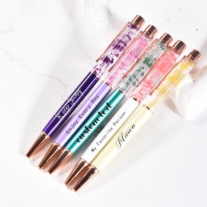 May include: A selection of colourful pens featuring floral designs encased within clear barrels. The pens have gold-coloured accents and include text such as "Keep Young", "Enjoy Every Day", "custom text", and "My Favorite Person Mason".