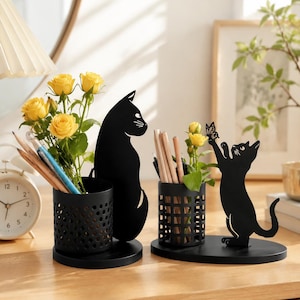 Könnte beinhalten: Zwei Schreibtisch-Organizer aus schwarzem Metall mit Katzensilhouetten. Einer hat einen perforierten zylindrischen Becher mit gelben Rosen und Stiften. Der andere hat einen gitterförmigen Becher und eine Katze, die nach einem Schmetterling greift.