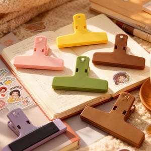 May include: A collection of colorful plastic bag clips in shades of pink, yellow, green, brown, and purple. The clips are arranged on an open notebook with stickers. The clips are designed to seal bags and keep contents fresh.