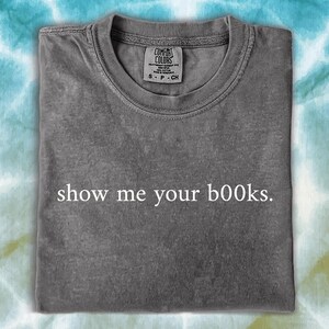 Pode incluir: Uma t-shirt cinzenta com a frase "show me your books". A t-shirt está dobrada e tem uma etiqueta no canto superior direito. O fundo é um padrão tie-dye em tons de azul e verde.