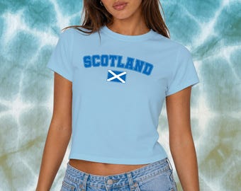 Puede incluir: Top corto azul claro con la palabra "SCOTLAND" en letras azules arqueadas. Una bandera escocesa blanca y azul está centrada debajo. El top se combina con vaqueros azules.