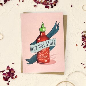 May include: A pink greeting card with a blue and white banner that says "Hey Hot Stuff". The banner is wrapped around a red hot sauce bottle with a green cap.