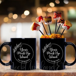 May include: Two black coffee mugs with white text that reads "Your Mock Up Island" and "Your Design Here". The mugs are on a wooden surface with a blurred background of lights and a bouquet of orange flowers with chocolate covered marshmallows on sticks.