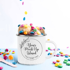 May include: White enamel mug with a black and white design that says "Your Mock Up Island" and "Your Design Here..." filled with cookie dough and colorful candy sprinkles. The mug is on a light blue surface with more sprinkles scattered around it.
