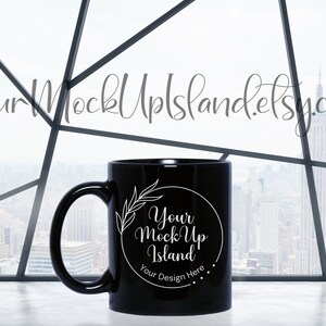 Może przedstawiać: Czarny kubek ceramiczny z białym wzorem, na kt&oacute;rym widnieje napis "Your Mock Up Island" i "Your Design Here". Kubek stoi na białym blacie przed oknem z widokiem na miasto.
