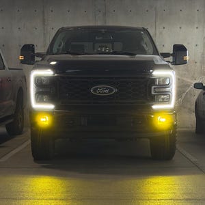 May include: A black Ford truck with bright white headlights and yellow fog lights is parked in a garage. The truck's front grill is visible, and the Ford logo is centered above it. The ground is illuminated with a yellow glow.