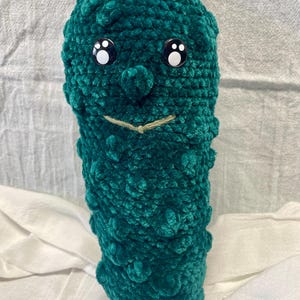 May include: A green crocheted plush toy shaped like a pickle with a smiling face and black button eyes.