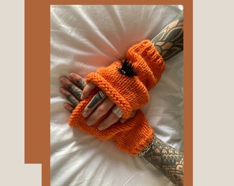 May include: A pair of orange knitted fingerless gloves with a black feather detail. The gloves are worn on a person's hands with tattoos.
