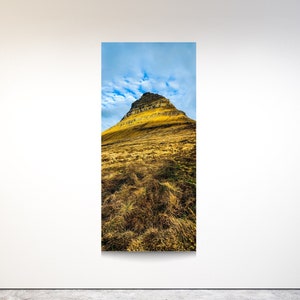 May include: A vertical landscape photograph of a mountain with a pointed peak. The mountain is golden yellow and brown, with patches of green grass at the base. The sky is blue with white clouds.