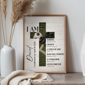 May include: Framed artwork with a wooden cross design. The cross incorporates a soccer ball, cleats, and the words "I AM." Text includes "Daniel Confirmed on April 14, 2024" and Bible verses. The background has a white wood plank design.
