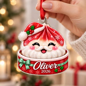 May include: A clear acrylic Christmas ornament shaped like a dumpling wearing a red Santa hat with white trim and holly berries. The base features a red band with a green bow, the name Oliver, and the year 2026, suspended by a red ribbon.