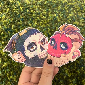 May include: Two skull-shaped stickers. One has a black hood and a cream-coloured face with brown eyes. The other has a red face with blue eyes and a tan outline. Both have glitter accents.