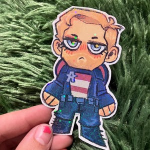 Pu&ograve; includere: Un adesivo con un'illustrazione a cartoni animati di Capitan America. Il personaggio indossa un'uniforme blu con il petto a strisce bianche e rosse. L'adesivo ha accenti glitterati.