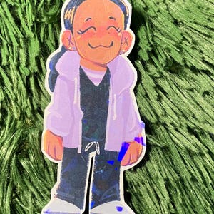K&ouml;nnte beinhalten: Ein Aufkleber mit einer Cartoon-Illustration einer Person, die eine lila Kapuzenjacke, ein schwarzes Hemd und eine schwarze Hose tr&auml;gt. Die Person hat dunkles Haar und l&auml;chelt. Der Aufkleber hat einen holografischen Effekt.
