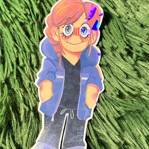K&ouml;nnte beinhalten: Ein Aufkleber mit einer Cartoon-Illustration einer Person mit roten Haaren, blauen Augen und einer blauen Jacke. Die Figur tr&auml;gt ein schwarzes Hemd und eine graue Hose. Der Aufkleber hat ein holografisches Finish.