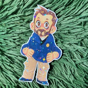 May include: A sticker depicting a cartoon man with a beard, wearing a blue jacket with star accents and tan trousers. The sticker has glitter accents and a white border. The man has rosy cheeks and large eyes.