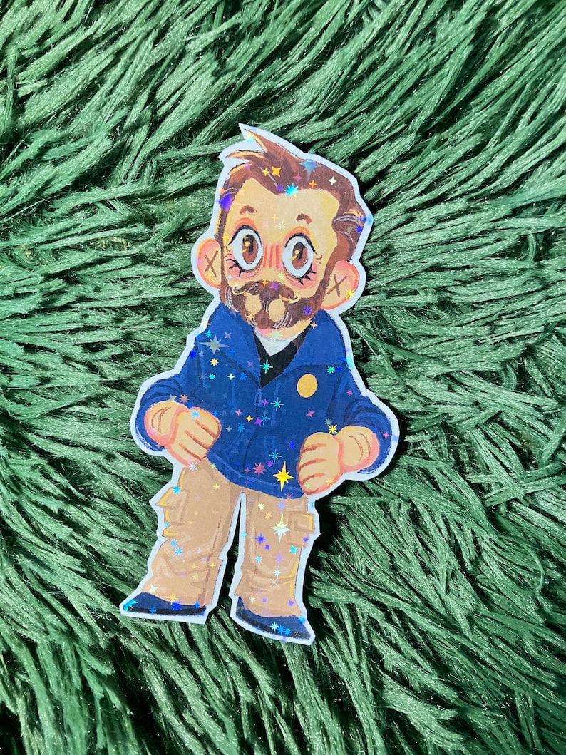 K&ouml;nnte beinhalten: Ein Aufkleber mit einer Cartoon-Illustration eines Mannes mit Bart, der eine blaue Jacke mit Sternakzenten und eine beige Hose tr&auml;gt. Der Aufkleber hat Glitzerakzente und einen wei&szlig;en Rand. Der Mann hat rosige Wangen und gro&szlig;e Augen.