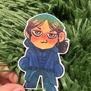 Pu&ograve; includere: Un adesivo raffigurante un personaggio dei cartoni animati in un completo blu con dettagli glitterati. Il personaggio ha i capelli castani, gli occhi arancioni e un'espressione neutra. L'adesivo &egrave; su una superficie verde e sfocata.