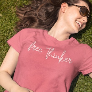May include: A woman wearing a pink t-shirt with the text "free thinker" in white script. She is lying on her back in a grassy field and smiling.