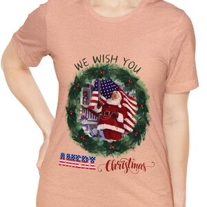 May include: Peach-coloured t-shirt with a festive design. The graphic features a wreath, Santa Claus holding an American flag, and the text "We Wish You Merry Christmas". The design incorporates red, white, and blue colours.