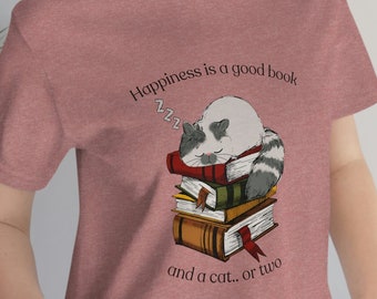 Op de afbeelding: Een roze T-shirt met een afbeelding van een wasbeer die slaapt op een stapel boeken. De tekst op het shirt luidt "Happiness is a good book and a cat... or two."