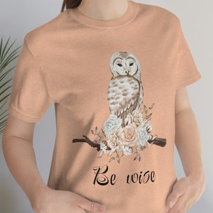 May include: A peach-colored t-shirt with a white owl perched on a branch with pink and white flowers. The text "Be wise" is printed below the owl.