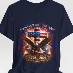 May include: Navy blue t-shirt featuring a patriotic design. The graphic includes a cross with the American flag, an eagle, and the text "Faith, Freedom, 250 Years" and "1776-2026". The design is surrounded by fireworks.