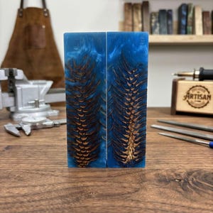 May include: Two rectangular blocks of blue resin with embedded brown pine cone details. The resin has a swirling, marbled effect. The blocks are displayed on a wooden surface, showcasing the artisan craftsmanship.
