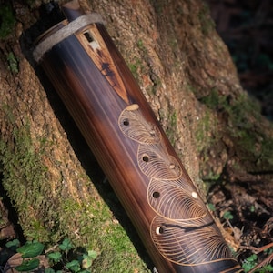 Puede incluir: Didgeridoo artesanal de bambú marrón oscuro con intrincados diseños grabados. El instrumento presenta una forma cilíndrica, patrones circulares decorativos y un acabado natural. Se apoya contra un tronco de árbol cubierto de musgo.